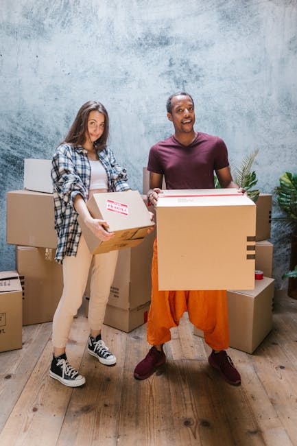 Two individuals engaged in a home relocation process inside a room with a textured gray wall and wooden flooring, surrounded by multiple cardboard boxes of various sizes. The woman on the left, dressed in casual clothing including a plaid shirt, beige pants, and sneakers, is holding a medium-sized box with a red and white 'Fragile' label, preparing to load or move it. The man on the right, wearing a maroon t-shirt, orange pants, and maroon shoes, is holding a larger cardboard box and smiling, suggesting an active packing or lifting action as part of furniture transport or packing and moving activities. Behind them, additional packed boxes are stacked, and a green potted plant is visible in the background, indicating an organized interior setup for a domestic moving or furniture transport scenario, which the professional team at Movers Knightsbridge might handle as part of their house removals services.