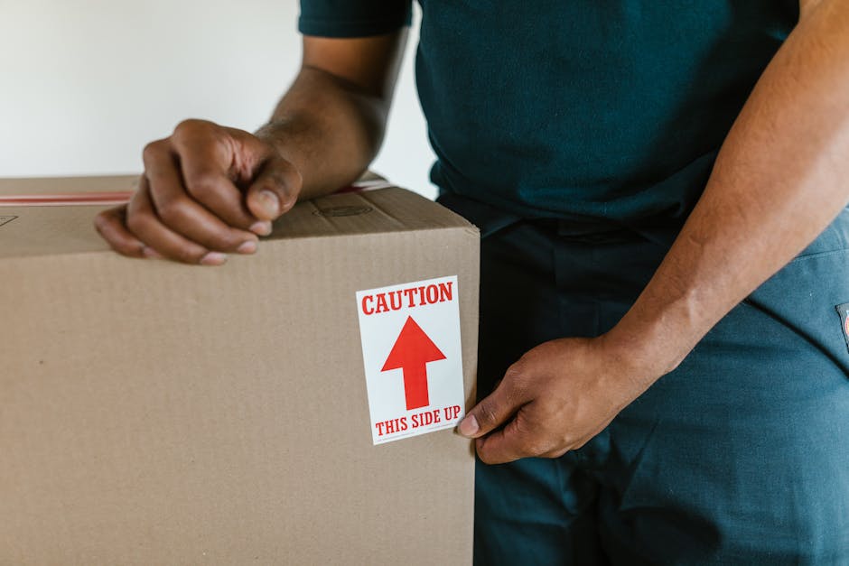 A person wearing a dark green shirt and blue trousers is holding a large cardboard box with a red and white caution label that reads 'Caution This Side Up' and features an upward-pointing arrow, indicating careful handling during home relocation. The individual is gripping the box firmly, with one hand positioned on the top edge and the other on the side, inside a room with neutral-colored walls. The box appears to contain fragile items, evident from the protective plastic wrapping and packaging materials visible around it. The scene suggests an active loading process, likely part of furniture transport and packing during a house removal service. The background shows part of a doorway or passage, with the focus on the careful handling of the box, supported by professional movers such as Movers Knightsbridge, who specialise in timely and secure moving logistics for residential relocations.
