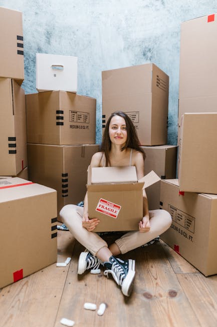 Two individuals engaged in a home relocation process inside a room with a textured gray wall and wooden flooring, surrounded by multiple cardboard boxes of various sizes. The woman on the left, dressed in casual clothing including a plaid shirt, beige pants, and sneakers, is holding a medium-sized box with a red and white 'Fragile' label, preparing to load or move it. The man on the right, wearing a maroon t-shirt, orange pants, and maroon shoes, is holding a larger cardboard box and smiling, suggesting an active packing or lifting action as part of furniture transport or packing and moving activities. Behind them, additional packed boxes are stacked, and a green potted plant is visible in the background, indicating an organized interior setup for a domestic moving or furniture transport scenario, which the professional team at Movers Knightsbridge might handle as part of their house removals services.
