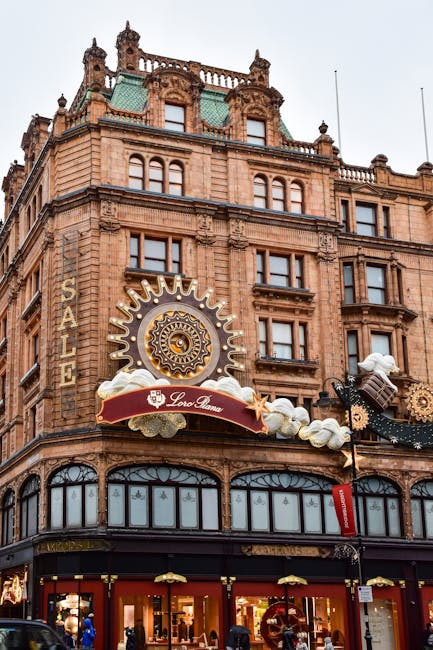 A tall, historic multi-storey building with architectural detailing and large windows at street level, displaying the sign 'Harrods' in vertical, illuminated letters on its corner façade. The buildin