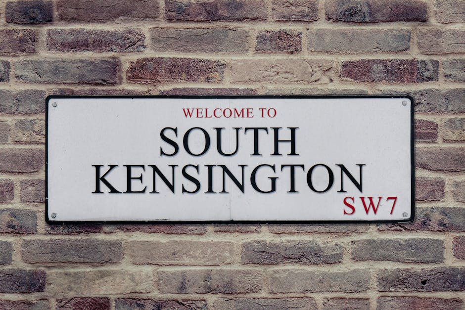 A rectangular white metal sign mounted on a brick wall, displaying the text 'WELCOME TO SOUTH KENSINGTON SW7' in black and red lettering. The sign is positioned outdoors, attached with visible screws at each corner, and is set against a background of reddish-brown bricks with a slightly textured surface. The image depicts a typical street or building façade in the South Kensington area, relevant for house removals and relocation services offered by Movers Knightsbridge, illustrating the initial point of contact or entry in a home relocation process involving furniture transport and packing and moving activities.