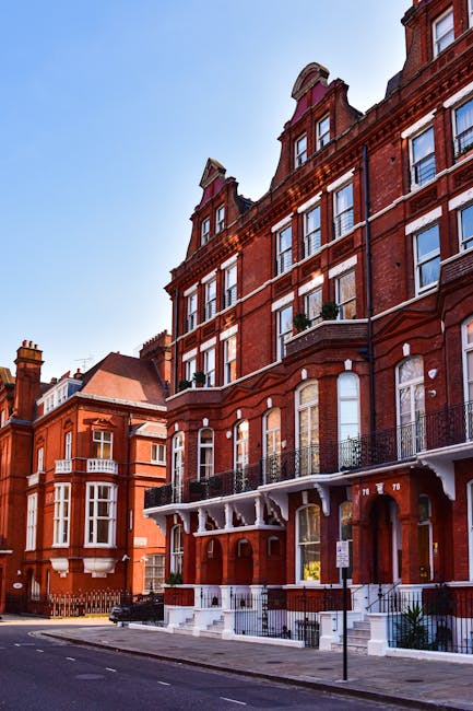 A row of red-brick Victorian terraced houses on a tree-lined street in Knightsbridge, with the artistically designed upper floors featuring large sash windows and decorative stonework. In the foreground, a white mobile staircase with wheels is positioned on the pavement, connected to an open internal doorway of one of the buildings. Inside the doorway, a moving team from Movers Knightsbridge appears to be engaged in the home relocation process, with furniture and cardboard boxes stacked nearby, some covered with protective blankets and plastic wrap. A black trolley is loaded with moving supplies, and the loading process is underway as part of the furniture transport in preparation for a house move. Bright daylight casts shadows on the building facades, emphasizing the architectural details, while the street remains quiet with minimal traffic and no pedestrians present, reflecting a professional moving operation at a residential property in Kensington and Chelsea.