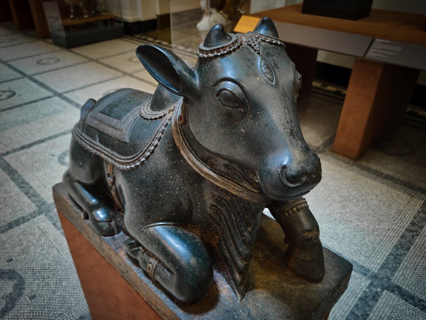 A detailed bronze statue of a cow, positioned indoors on a wooden platform in a room with a patterned carpet. The sculpture features intricate decorative elements, including a saddle-like covering on its back and ornate adornments around the head and neck. The cow's ears are perked, and it has a calm expression. In the background, there is a wooden table with a drawer, some paper or booklet, and part of a plastic-wrapped object. The lighting appears to be natural, with soft shadows highlighting the textured surface of the statue. This image may relate to home decoration or cultural artifacts within a house, which could be relevant for a relocation or house removals service like Movers Knightsbridge during packing and moving procedures.