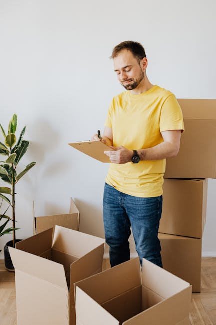 A man with short dark hair and a beard, wearing a yellow t-shirt and blue jeans, stands inside a well-lit room with white walls, surrounded by open cardboard boxes and packing materials. He is holding a clipboard and pen, appearing to review or check items for a home relocation or furniture transport process by Movers Knightsbridge. Behind him, there are large stacked cardboard boxes, some partially open, indicating active packing or unpacking. To his left, a green houseplant in a pot adds a touch of decor to the space. The scene captures the preparation phase of house removals, with a focus on organized packing activities in a high-value property setting. This visual emphasizes the detailed planning and logistics involved in the professional moving service for high-value properties, aligning with the content about Knightsbridge removals checklist for high-value properties.