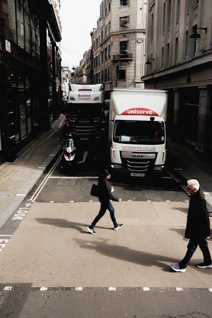 A street scene captured from an elevated perspective showing a moving van parked partially on the pavement and street, with the company logo 'Uniserve' and website visible on the side and front of the van. The vehicle is positioned at the edge of a narrow road lined with tall, multi-storey buildings on both sides, typical of central London architecture. In front of the van, there is a motorbike parked parallel to the curb on a designated parking lane. Pedestrians are crossing the street: a woman dressed in dark clothing carrying a shoulder bag and a man with white hair wearing a dark coat, both walking past the parked vehicles. The environment suggests a busy urban area suitable for household or commercial relocations, where moving companies like Movers Knightsbridge arrange for the careful transportation of furniture and boxes during home relocation or moving services, including loading and unloading activities around tight urban spaces.