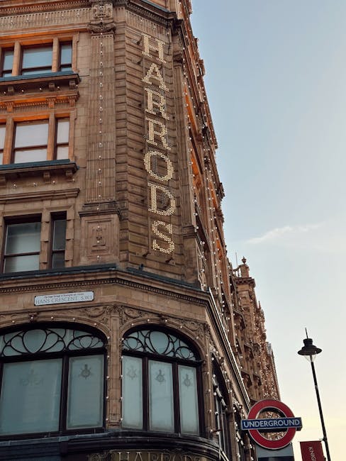 Photograph of a historic multi-storey building with intricate stone detailing and large arched windows on the ground floor, located on a city street. The building has a prominent vertical sign spelling 'FAYRERODS' mounted along its corner facade, with white lighting fixtures highlighting the sign against the weathered stone exterior. Above, the sky is clear with minimal clouds, and the street scene includes a traditional black street lamp. A round London Underground sign with a blue bar and red circle is visible near the corner, indicating the proximity to a tube station. The scene suggests an urban environment suitable for home relocation activities, and the building's exterior and street elements are consistent with a busy city area where professional removals services like those from Movers Knightsbridge may operate, supporting the themes of packing, furniture transport, and loading processes associated with house removals.