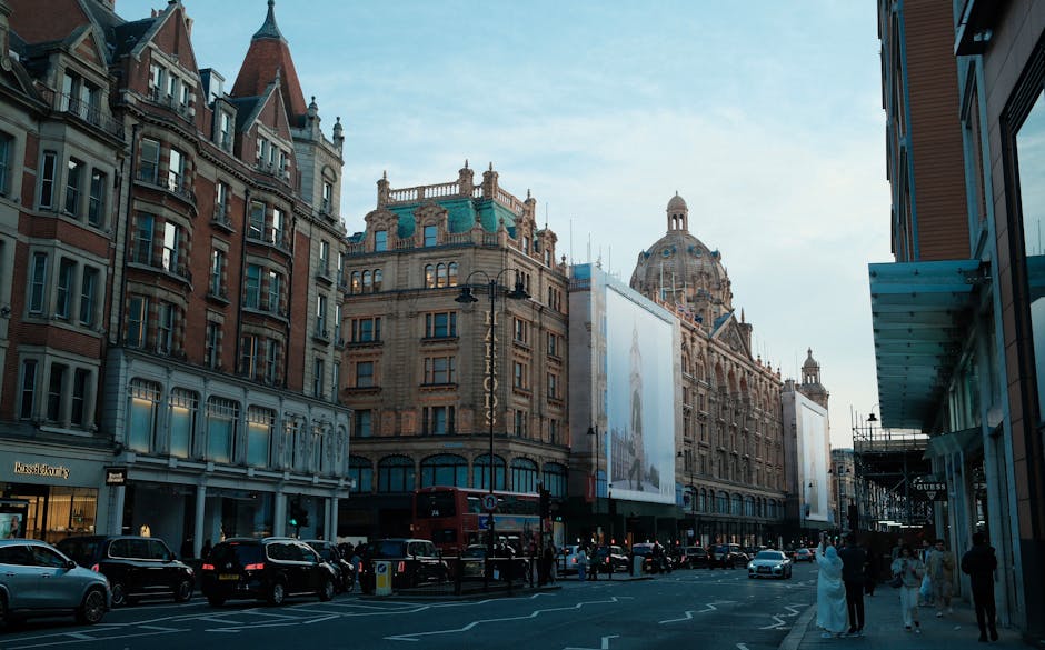 A street scene captured from an elevated perspective showing a moving van parked partially on the pavement and street, with the company logo 'Uniserve' and website visible on the side and front of the van. The vehicle is positioned at the edge of a narrow road lined with tall, multi-storey buildings on both sides, typical of central London architecture. In front of the van, there is a motorbike parked parallel to the curb on a designated parking lane. Pedestrians are crossing the street: a woman dressed in dark clothing carrying a shoulder bag and a man with white hair wearing a dark coat, both walking past the parked vehicles. The environment suggests a busy urban area suitable for household or commercial relocations, where moving companies like Movers Knightsbridge arrange for the careful transportation of furniture and boxes during home relocation or moving services, including loading and unloading activities around tight urban spaces.
