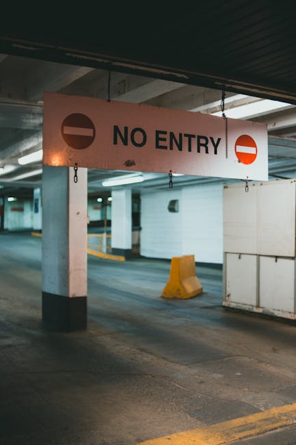 A temporary parking area within an underground or multi-story car park shows a prominent 'No Entry' sign hanging from the ceiling, supported by metal chains attached to concrete pillars. The sign has a white background with black text and red circular prohibition symbols on each end. The area features concrete flooring with painted yellow and white lines marking designated parking spaces. Nearby, a bright yellow wheel stop barrier is positioned on the pavement, and part of a white wall and structural columns are visible in the background. Under the ceiling, there are overhead lighting fixtures providing illumination. This setting indicates a restricted zone where vehicle entry is prohibited, reflecting typical conditions during home relocation or furniture transport processes managed by specialists such as Movers Knightsbridge, with a focus on logistical operations within limited or controlled environment areas.