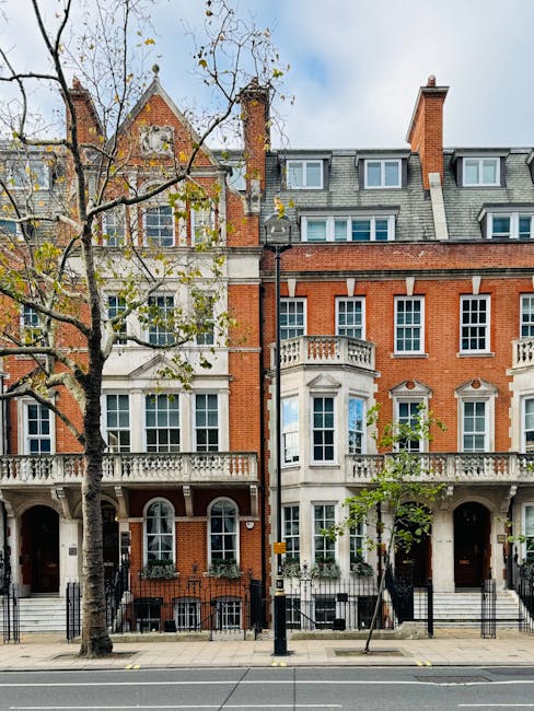 A row of elegant, multi-storey residential buildings on a tree-lined street with a paved sidewalk, showcasing a mix of red brick and white façade exteriors, featuring large sash windows, decorative railings, and balconies. The buildings are used as part of a home relocation and furniture transport process, with a moving van from Movers Knightsbridge visible on the street, adjacent to the buildings, ready for loading or unloading. Some windows are open, revealing interior furnishings, while the street is calm with a few trees showing early autumn foliage, and the overall scene reflects a typical urban environment suited for professional removals and packing and moving services in Knightsbridge.