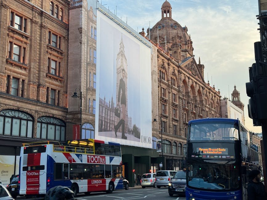 A busy street scene in Knightsbridge featuring historic and ornate building facades with intricate stonework and large windows. Prominently visible is a large vertical billboard advertising Dior, mounted on the building exterior. In the foreground, a double-decker bus with a digital destination display and a red and white color scheme is stopped alongside other vehicles, including a smaller white van and several cars. The street is bustling with movement during daylight hours, and the surrounding environment suggests a high-end shopping district with a mix of modern and historic architecture, typical of a home relocation or furniture transport context where removals services by Movers Knightsbridge might operate.