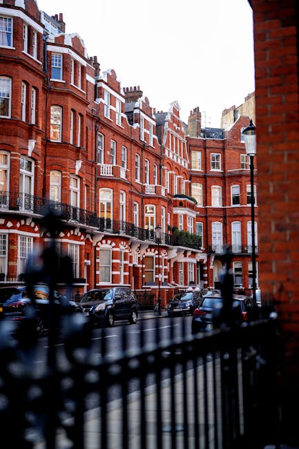 A row of elegant, multi-storey residential buildings on a tree-lined street with a paved sidewalk, showcasing a mix of red brick and white façade exteriors, featuring large sash windows, decorative railings, and balconies. The buildings are used as part of a home relocation and furniture transport process, with a moving van from Movers Knightsbridge visible on the street, adjacent to the buildings, ready for loading or unloading. Some windows are open, revealing interior furnishings, while the street is calm with a few trees showing early autumn foliage, and the overall scene reflects a typical urban environment suited for professional removals and packing and moving services in Knightsbridge.